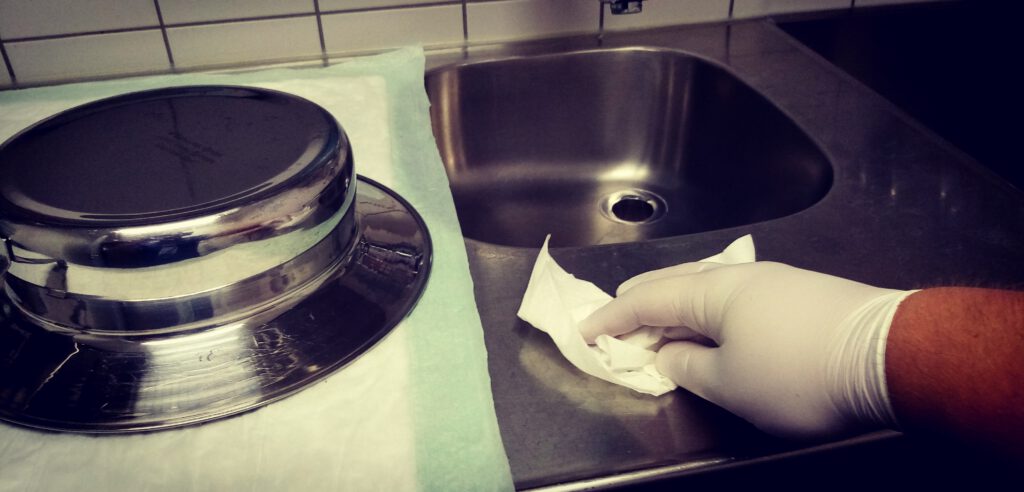 Ein Unreiner Arbeitsraum in einem Krankenhaus. Links liegt umgedreht eine Bettschüssel, rechts poliert eine Hand die Arbeitsfläche mit einem Tuch