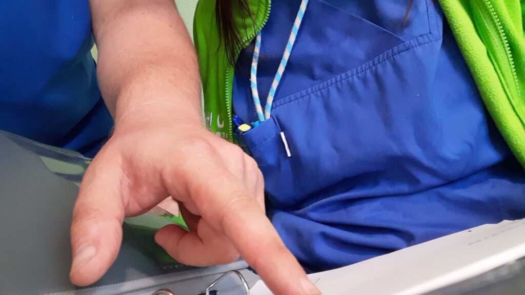 Zwei Pflegende sitzen vor einem Ordner. Man sieht nur die Oberkörper und eine Hand, die auf den Ordner zeigt.